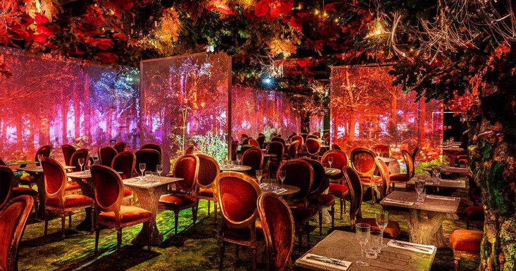 Une vue de la salle à manger immersive du restaurant Wonderwoods à Angers, décorée de chaises en velours rouge et de murs projetant une forêt féerique, pour un top des restaurants d'Angers.